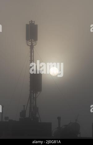 Kommunikationsturm auf einem Gebäude mit 4G 5G Antenne bei Sonnenaufgang im Nebel. Stockfoto