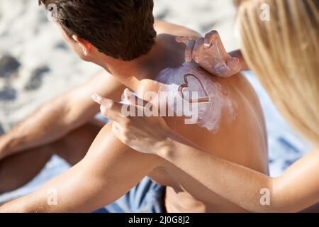 Sie zeigt ihre Liebe wo immer sie kann. Eine junge Frau, die eine Herzform mit Sonnencreme auf dem Rücken ihres Freundes macht. Stockfoto