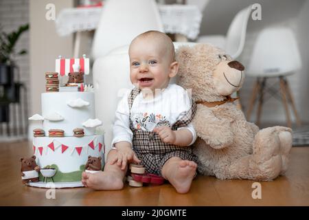 Lächelnder kleiner Junge in weißem T-Shirt und kariertem Overall, der auf dem Boden neben zweistufigem Geburtstagskuchen und Teddybär sitzt. Stockfoto