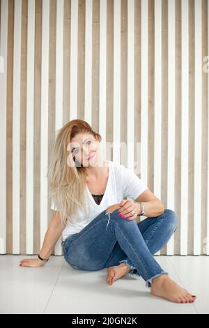 Lächelnde Frau mittleren Alters mit langen, hellen Haaren in weißem T-Shirt und blau zerrissenen Jeans, die mit gekreuzten Beinen sitzen. Stockfoto