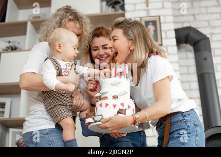 Drei lächelnde, wunderschöne Frauen mittleren Alters und junge Frauen gratulieren dem süßen, kleinen einjährigen Jungen, der den Makronen-Kuchen berührt. Stockfoto