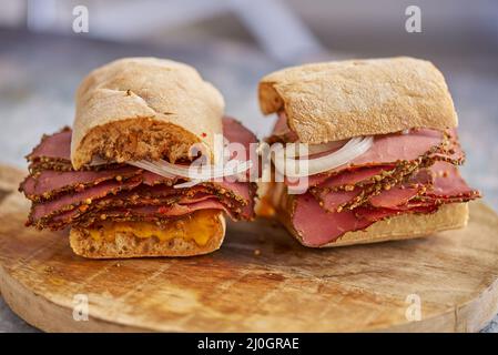 Dünn geschnittener Pastrami-Sand, der sich mit köstlichen Speisen auf Baguette-Brot mit Käsekäse, Zwiebeln trifft Stockfoto