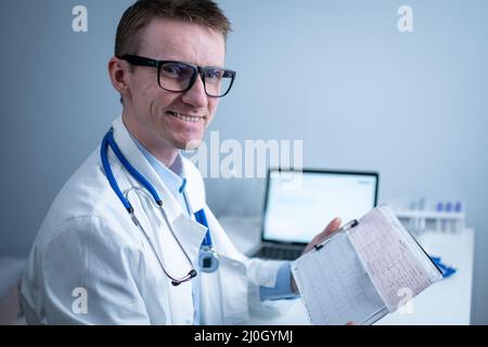 Junger, gutaussehender Arzt mit Brille und weißem Laborkittel untersucht die Ergebnisse der elektrokardiographischen Untersuchung des Patientenherzens in Stockfoto