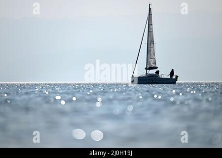 Langenargen, Deutschland. 19. März 2022. Vor Langenargen segelt ein Boot auf dem Bodensee, während im Vordergrund die Sonne im Wasser glitzert. Quelle: Felix Kästle/dpa/Alamy Live News Stockfoto