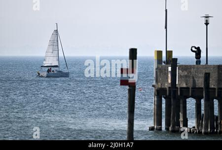 Langenargen, Deutschland. 19. März 2022. Ein Boot fährt am Bodensee vor Langenargen, während eine Frau die Szene vom Pier aus fotografiert. Quelle: Felix Kästle/dpa/Alamy Live News Stockfoto