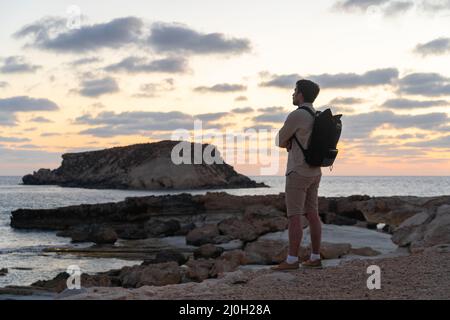 Blick auf den Sonnenuntergang. Mann mit Rucksack auf Felsen mit wunderschönem Blick auf die Insel Yeronisos nahe der Küste von Agios Georgios Pegeias. Guy en Stockfoto