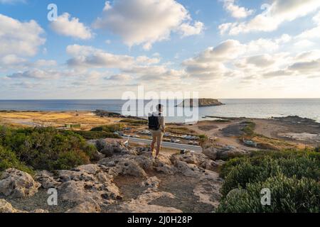 Blick auf den Sonnenuntergang. Mann mit Rucksack auf Felsen mit wunderschönem Blick auf die Insel Yeronisos nahe der Küste von Agios Georgios Pegeias. Guy en Stockfoto