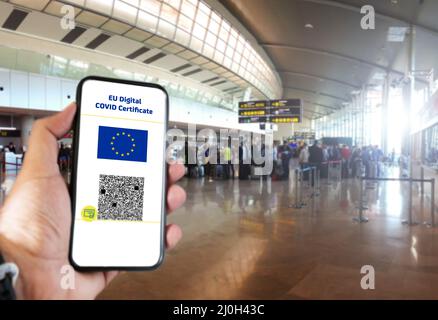 Digitales EU-COVID-Zertifikat mit dem QR-Code auf dem Bildschirm eines Mobiltelefons, das von einer Hand gehalten wird, wobei der Flughafen im Hintergrund unscharf ist Stockfoto