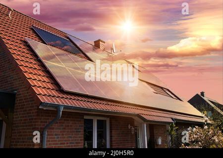 Fotocollage von Solarzellen, Photovoltaik auf dem roten Dach eines Hauses und einem schönen Himmel mit untergehenden Sonne. Alternative Stromversorgung. Konz Stockfoto