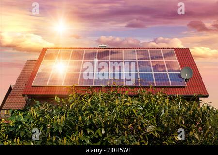 Fotocollage von Solarzellen, Photovoltaik auf dem roten Dach eines Hauses und einem schönen Himmel mit untergehenden Sonne. Alternative Stromversorgung. Konz Stockfoto