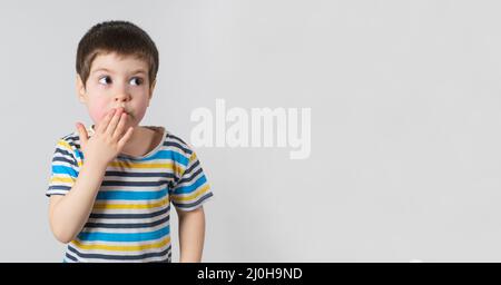 Ein hübscher Junge von 4 Jahren in einem gestreiften T-Shirt bedeckt seinen Mund mit seiner Hand und schaut zur Seite, ein Banner mit Platz für Text Stockfoto