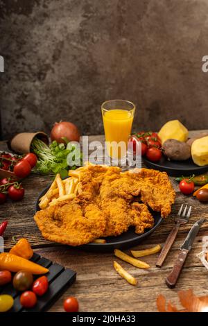 Goldfarbenes Schweinebraten mit gebratenen Pommes auf einem schwarzen Teller mit gemütl. Beilage auf dem Holztisch Stockfoto