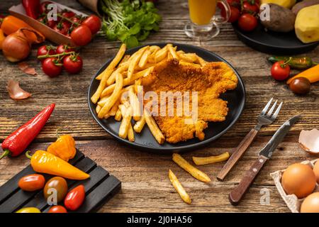 Goldfarbenes Schweinebraten mit gebratenen Pommes auf einem schwarzen Teller mit gemütl. Beilage auf dem Holztisch Stockfoto