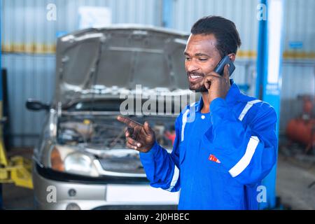 Lächelnder Automechaniker, der mit dem Kunden vor einem kaputten Auto in der Garage telefoniert - Konzept der Entspannung, Pause und Kundenservice Stockfoto