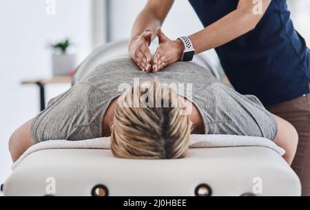 Ihr Rücken braucht all die Erleichterung, die er bekommen kann. Aufnahme einer reifen Frau, die sich mit dem Gesicht nach unten legt und von einem Physiotherapeuten in einer Klinik massiert wird. Stockfoto