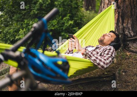 Der Mann auf einer Fahrradtour beim Camping am See entspannt sich in einer grünen Hängematte, während er Musik hört. Aktive Erholung Thema in der Natur. Stockfoto