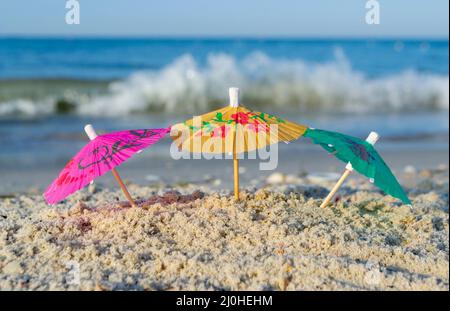 Drei kleine Papier-Cocktail-Schirme stehen im Sand am Sandstrand aus nächster Nähe. Stockfoto