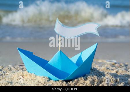Blaues Papierboot auf einem Sandstrand in der Nähe des Meeres an einem sonnigen Sommertag. Stockfoto