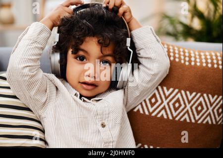 Dieses Lied lässt mich meine Coolness verlieren. Porträt eines entzückenden kleinen Jungen, der Musik über Kopfhörer hört, während er zu Hause auf einem Sofa sitzt. Stockfoto