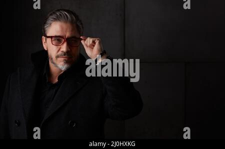 Porträt eines älteren Mannes im Jahr 50s mit grauen Haaren und Brille vor dunklem Hintergrund. Tragen von Mantel, Kopierraum. Stockfoto