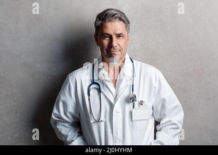 Porträt eines vertrauenswürdigen älteren, selbstbewussten Arztes mit grauen Haaren in einem weißen Laborkittel, der an einer grauen Wand steht und lächelt. Speicherplatz kopieren. Stockfoto