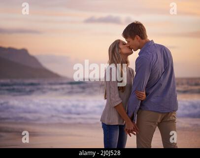 Jeder Kuss fühlt sich an wie unser erster. Aufnahme eines jungen Paares, das bei Sonnenuntergang einen romantischen Kuss am Strand genießt. Stockfoto