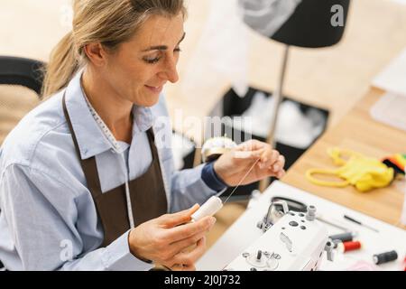 Junge Schneiderin Frau näht Kleidung auf Arbeitstisch. Lächelnde Näherin und ihre Hand in der Werkstatt. Fokus auf Naht auf Tisch Stockfoto