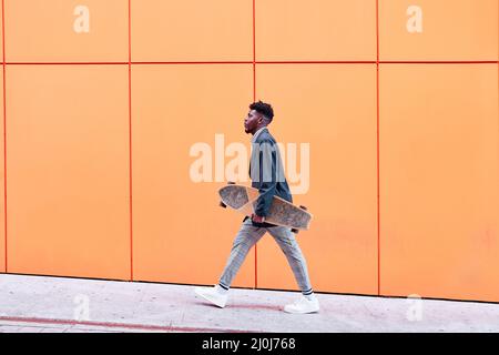 Seitenansicht eines jungen Mannes mit Blazer und Skateboard, der vor orangefarbenem Hintergrund die Straße entlang läuft. Stockfoto