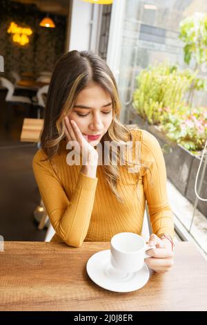 An einem Tisch sitzend eine schöne, junge, lateinische Frau mit langen blonden Haaren und schönem Make-up, Restaurant-Interieur am Tag, heiße Getränke Stockfoto