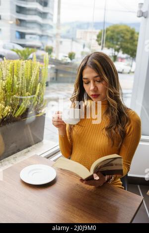 Frühstück mit einer Tasse mit einem heißen Getränk und Lesen eines Buches, junge lateinische Frau mit langen blonden Haaren, Lifestyle und intellektuelles Hobby, Kaffeepause Stockfoto