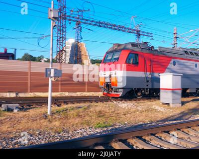 Die Vorderseite eines Zuges, der an einem sonnigen Tag vor der Kulisse eines Zauns und eines Gebäudes entlang der Schienen rast Stockfoto