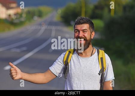 Reisen mit Autostop, mit Sommerausflug. Auto-Stopp-Fahrt. Mann mit strengem Gesicht und Bart, der durch Anhalter mit Straße im Hintergrund reist. Stockfoto