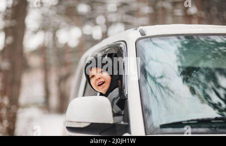 Aufgeregter kleiner Junge, der den Kopf aus dem Autofenster ragt, während er durch verschneiten Wald geht Stockfoto