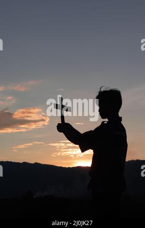 Silhouette eines Mannes, der das christliche Kreuz in den Händen hält, um im Hintergrund des Sonnenuntergangs für den Segen zu beten Stockfoto