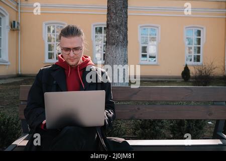 Junger Mann, Student, der online über einen Laptop lernt, sitzt auf einer Bank auf einem Campus. Stylish Hipster Kerl erfahrene Freiberufler arbeiten d Stockfoto