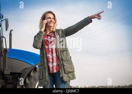 Eine junge attraktive Frau telefoniert vor einem Traktor auf einem Feld. Stockfoto