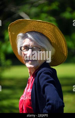 Eine hundertjährige Landfrau steht im Freien in ihrem roten karierten Hemd und Strohhut. Die Sonne scheint auf ihrem weißen Haar. Stockfoto