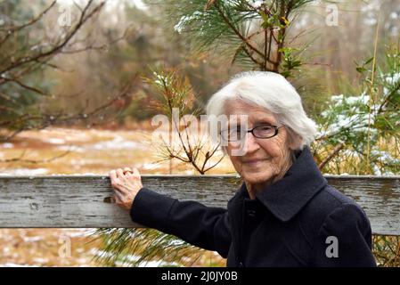 Die 100-jährige Frau hält an einem weißen, rustikalen Holzzaun inne. Schnee liegt auf Kiefernästen. Sie ist in einen grauen Wollmantel gekleidet. Stockfoto