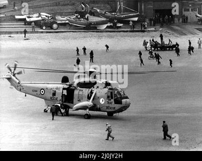 Ein RAF-Such- und Rettungsdienst Sea King von RAF Boulmer landet einen Mann am Strand, nachdem er vor Cullercoats aus dem Meer gezupft wurde. Im Hintergrund wird das umgekehrte Küstenrettungsfahrzeug an Land gezogen. Der Hubschrauber war an der doppelten Rettung bei starken Winden beteiligt gewesen, die mit dem Kentern eines Fischerbootes begonnen hatte, gefolgt von dem Rettungsboot, das dasselbe tat. Zwei Männer wurden vom Seegang gerettet, ein anderer Mann schaffte es, zurück zum Ufer zu schwimmen, wo ein vierter von Algen und Seil aus dem Boot gefangen ertrunken war. 22/02/1981. Stockfoto