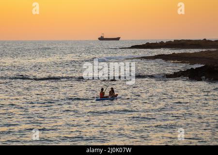 Zwei Mädchen sitzen auf dem Brett im ruhigen mittelmeer bei Sonnenuntergang in der Stadt paphos auf zypern. Silhouetten von 2 Mädchen Paddel Stockfoto