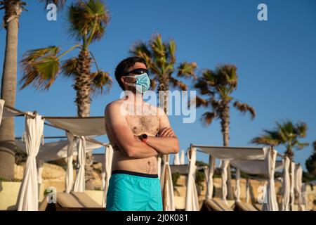 Ruhender Mann in einer Schutzmaske auf einem schmutzigen Strand allein vor dem Hintergrund von Palmen und Sonnenliegen auf dem Mittelmeer Stockfoto