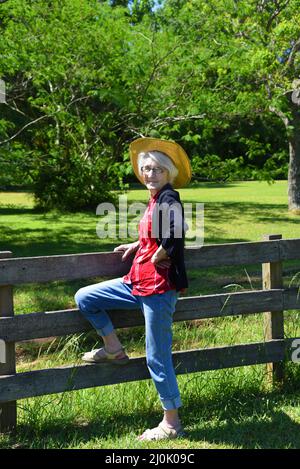 Ältere Frau ruht auf einem rustikalen, hölzernen Zaun. Sie webt ein rotes kariertes Hemd und Jeans. Ihr weißer Kopf ist mit einem Strohhut übersät. Sommer gr Stockfoto