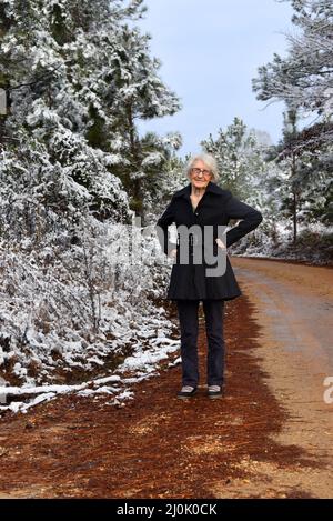 Die 100-jährige, ältere Frau genießt einen Spaziergang auf einer verlassenen Schotterstraße in South Arkansas. Schnee hängt an den Bäumen. Sie hat ihre Hände auf den Hüften, als ob sie defyi wäre Stockfoto