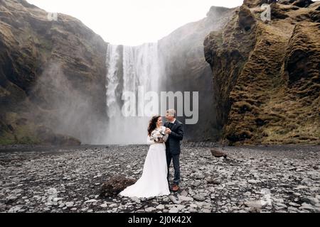 Hochzeitspaar in der Nähe des Skogafoss Wasserfalls. Reiseziel Island Hochzeit. Der Bräutigam umarmt die Braut an den Schultern, sie lachen zusammen Stockfoto