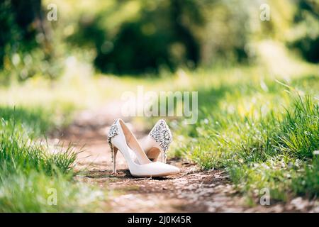 Weiße Hochzeitsschuhe der Braut, mit hohen Absätzen, verziert mit Schmucksteinen. Auf einem Waldweg zwischen dem grünen Gras Stockfoto