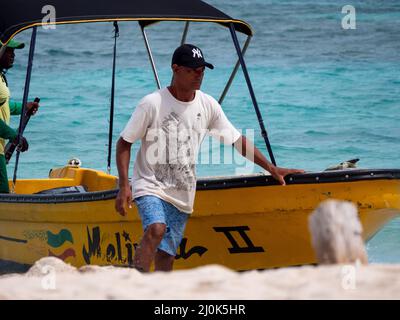 San Andrés, Kolumbien - November 17 2021: Brauner Mann in der Nähe eines gelben Bootes am Strand von Spratt Bight mit türkisfarbenem Meereshintergrund Stockfoto