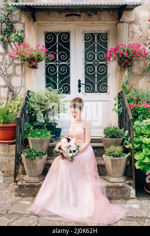 Schöne Braut in zartem Hochzeitskleid mit Brautstrauß sitzt auf der Veranda des Hauses in Perast Stockfoto