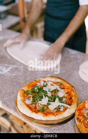 Fertige Pizza mit Käse und Rucola vor dem Hintergrund des Küchenchefs Pizza, rollt den Teig auf dem Tisch. Stockfoto