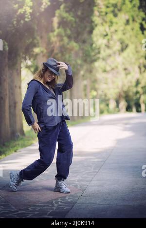 Lächelndes Mädchen in blauer Kleidung und einem Hut in tanzender Pose in einer Parkallee Stockfoto