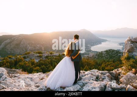 Eine Braut und ein Bräutigam stehen umarmt auf dem Berg Lovcen und Blick auf den Panoramablick auf die Bucht Von Kotor Stockfoto